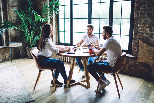 Full body of serious diverse colleagues in casual clothes sitting at wooden table and gathering business strategy in modern office - Fotografie, Obrázek