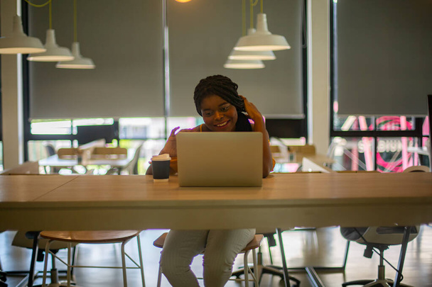 Νεαρή Αφροαμερικανή γυναίκα που εργάζεται χρησιμοποιώντας φορητό υπολογιστή συμπυκνωμένο και χαμογελαστό. - Φωτογραφία, εικόνα