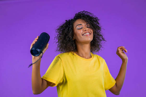 Mixed race woman listening to music by wireless portable speaker - modern sound system. African lady dancing, enjoying on purple studio background. She moves to the rhythm of music. - Fotografie, Obrázek