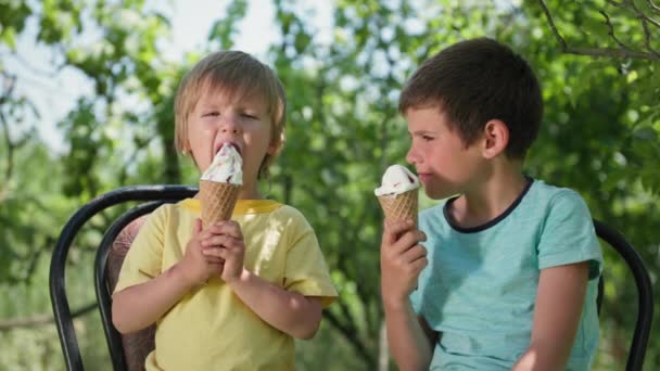 Şekerler, küçük çocuklar sıcak bir yaz gününde lezzetli dondurmalar yemekten hoşlanırlar yeşil ağaçların arkasında sandalyelerde otururlar. - Video, Çekim