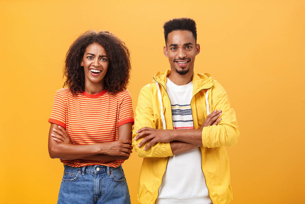 Portrait of surprised sarcastic african american woman with afro hairstyle standing with cute boyfriend crossing arms on chest laughing from scorn and fun over orange background - Foto, Imagem