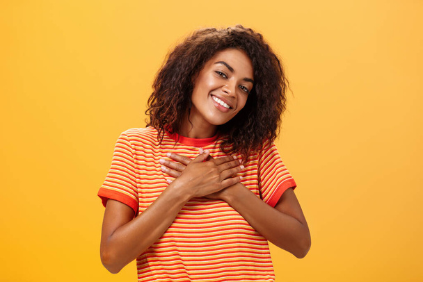 Pas-up záběr okouzlující smyslná žena tmavé pleti s kudrnatým účesem v trendy tričku drží dlaně na srdci hledá s potěšením milující úsměv pocit vděčný a potěšený přes oranžovou zeď - Fotografie, Obrázek