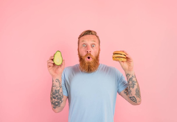 Amazed man with beard and tattoos is undecided if to eat an avocado or an hamburger - Photo, Image