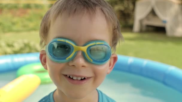 Zbliżenie twarz zabawny chłopiec gogle pozowanie na podwórku z nadmuchiwanym gumowym basenem - Materiał filmowy, wideo