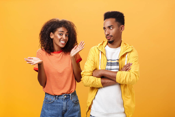 Guy thinks his friend weirdo making dumb thinks looking at cute girl with suspicious look crossing arms on chest raising eyebrow questioned while girlfriend saying sorry shrugging over orange wall - Photo, image