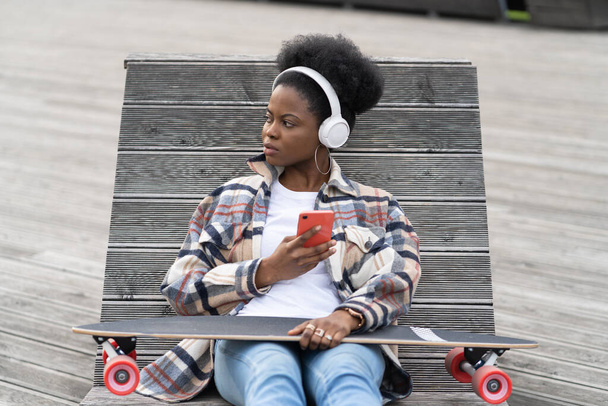 Joven africana skateboarder chat mujer en el teléfono inteligente. Chica patinadora negra en el espacio urbano con teléfono celular - Foto, imagen
