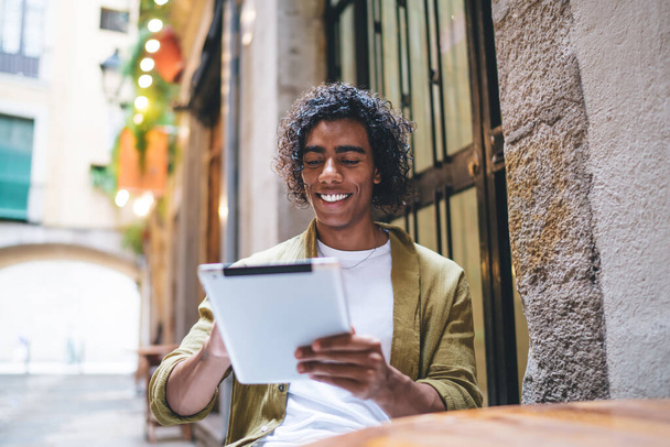 Low angle of positive young African American guy in casual clothes smiling while reading messages on tablet while resting in street cafe - Fotó, kép