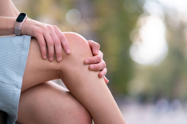 Primer plano de las manos de la mujer sosteniendo su rodilla con dolor sentado en el banco al aire libre. - Foto, imagen