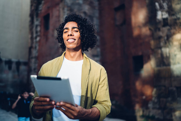 Low angle of positive confident young African American man in casual outfit smiling while communicating on tablet and enjoying sunny day against shabby brick building - Фото, зображення