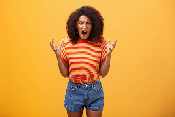 Pissed and outraged angry african american young female in stylish outfit raising palms gesturing and yelling losing temper arguing with cheating partner feeling fed up over orange background - Photo, Image