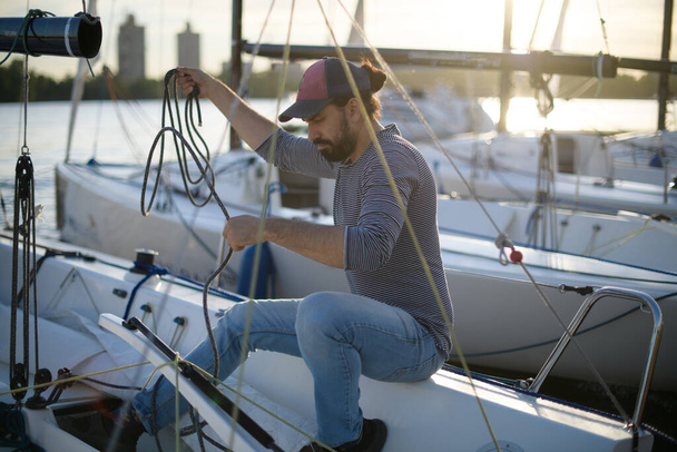 Preparation for the regatta at the yachting school at the marina with sailing yachts. A young male yachtsman prepares a moored sport yacht for sailing at sunset. - Foto, afbeelding