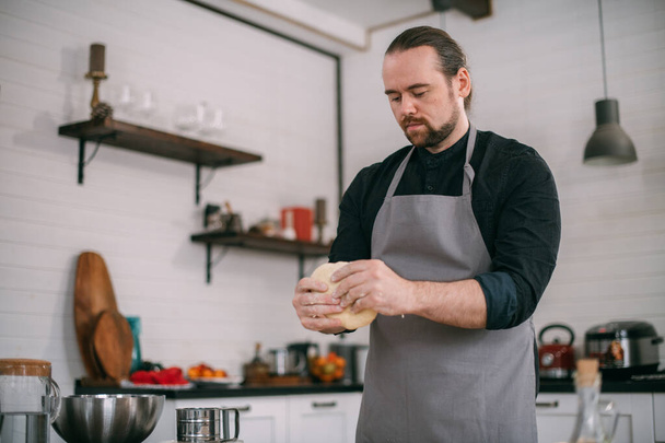 Un chef masculino prepara masa en casa en la cocina. Joven en un delantal en su cocina en la casa en la mesa con ingredientes para hacer masa - Foto, imagen