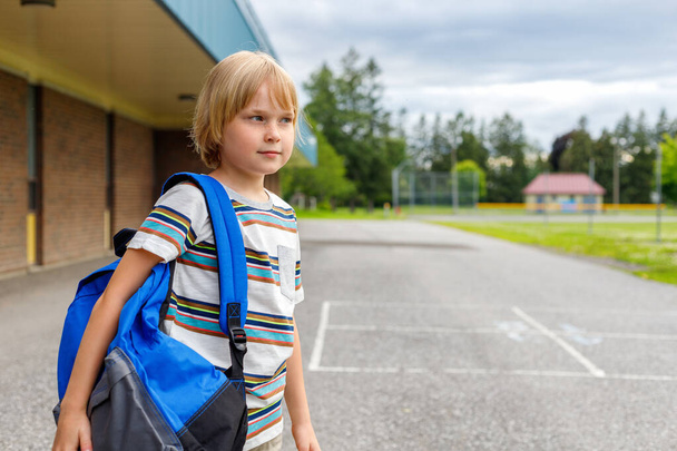 Beautiful child near school building at schoolyard. Little student carrying blue backpack, walking from school. Copy space for text. - Photo, Image