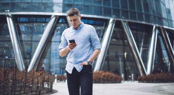 Focused young man in formal outfit surfing mobile phone while standing with hand in pocket against contemporary glass building on clear summer day - Foto, imagen