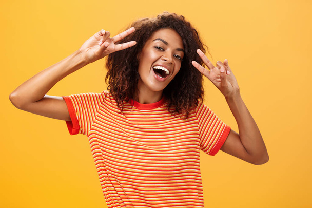 Nobody gonna spoil my perfect mood. Portrait of happy optimistic attractive dark-skinned female model with curly hairstyle tilting head smiling carefree showing peace or victory signs near face - Photo, Image