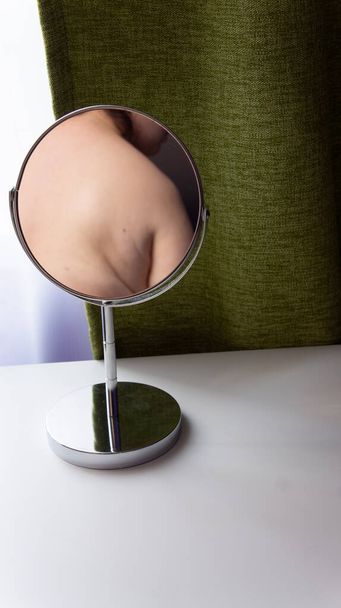 La figura di una donna grassa si riflette in uno specchio rotondo. Specchio si erge su un tavolo bianco su sfondo tende verdi - Foto, immagini