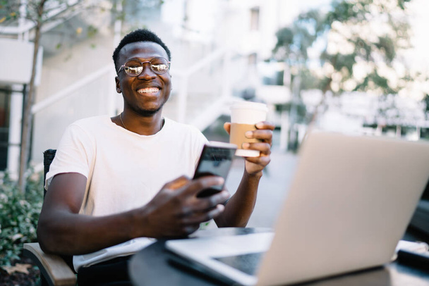 Smiling african american hipster guy in eyewear enjoying remote job on coffee terrace holding smartphone and coffee cup, positive dark skinned male freelancer sending text messages on cellular - Fotoğraf, Görsel