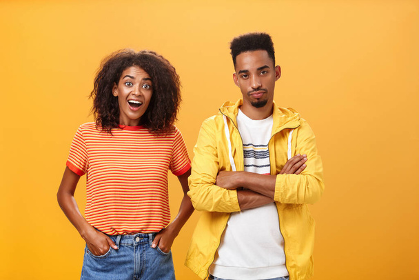 Two african american friends standing over orange background, girl thinks idea is awesome feeling excited and joyful while boyfriend being displeased and indifferent to unimpressive plan - Fotografie, Obrázek
