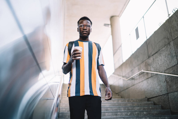 Half length portrait of trendy male tourist with caffeine beverage standing at urban stairs and looking at camera during solo travelling, African American hipster guy with takeaway coffee to go - Φωτογραφία, εικόνα