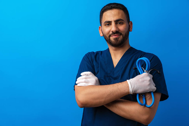 Handsome young male doctor in blue uniform with stethoscope against blue background - Photo, Image