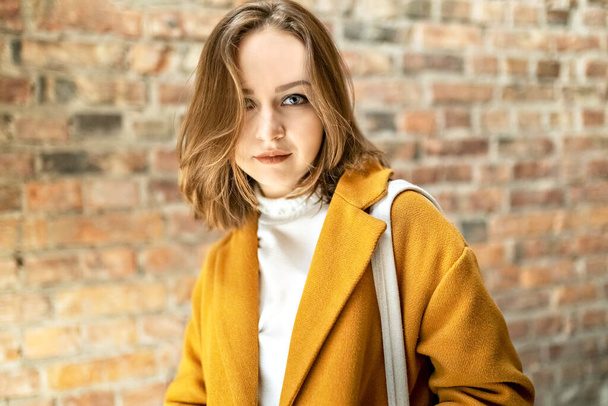 Портрет молодой женщины в ярко-желтом пальто на фоне кирпичной стены у окна. - Фото, изображение