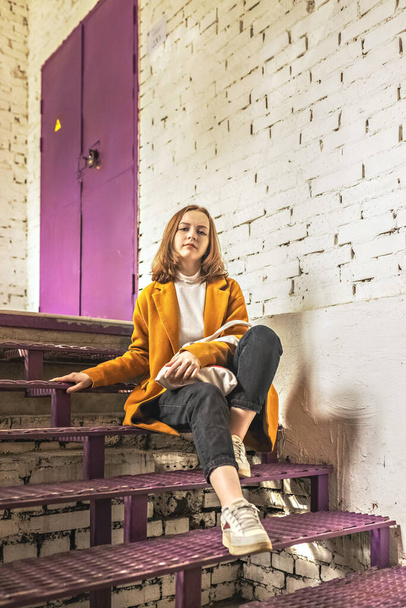 Портрет молодої дівчини-підлітка, що сидить на фіолетових сходах на білій цегляній стіні
. - Фото, зображення