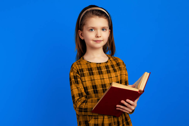 Serious attractive cute schoolgirl holding a book against blue background - Fotó, kép