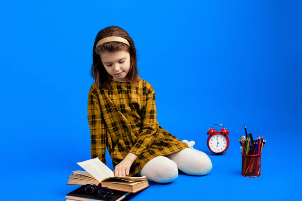 Cute schoolgirl kid reading book and sitting against blue background  - 写真・画像