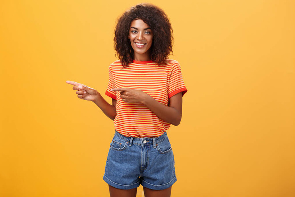 Portret uroczej, przyjacielsko wyglądającej afrykańsko-amerykańskiej współpracowniczki w luźnym stroju, wskazującej w lewo i uśmiechającej się szeroko pokazującej idealne miejsce na kopię dla klientów na pomarańczowym tle - Zdjęcie, obraz