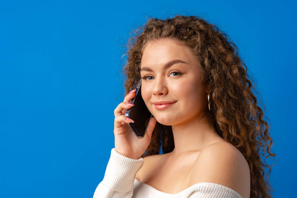 Young curly woman talking on the phone against blue background - Foto, afbeelding