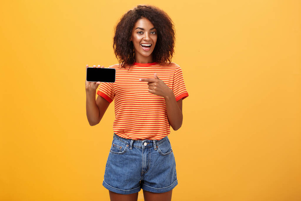 Girl showing picture in phone bragging about her cool vacation. Charming friendly and sociable african american woman with curly hair holding cellphone pointing at smartphone screen over orange wall - Fotografie, Obrázek