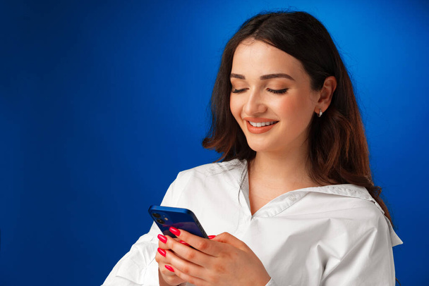 Smiling young woman in white shirt using her smartphone against blue background - Fotoğraf, Görsel