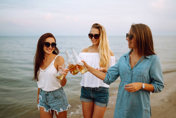 Three woman walking along the beach and drink beers.  Summer holidays, vacation, relax, picnic, party and lifestyle concept. - Fotó, kép