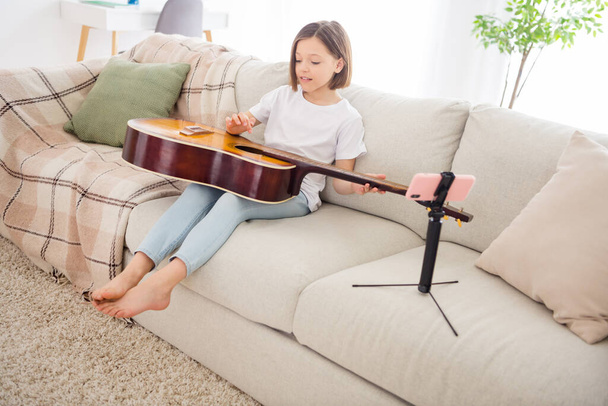 Photo of pretty cute schoolgirl dressed white t-shirt sitting coach playing guitar recording modern gadget indoors room home house - Foto, imagen