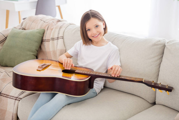 Photo of charming shiny school girl wear white t-shirt smiling sitting sofa learning playing guitar indoors house home room - Foto, Bild