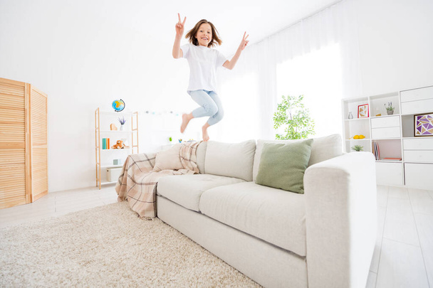 Photo of pretty funky small schoolgirl dressed white t-shirt showing v-signs jumping coach indoors room home house - Foto, imagen