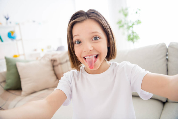 Photo of young cheerful little girl happy positive smile fooling grimace tongue-out make selfie indoors - Fotografie, Obrázek