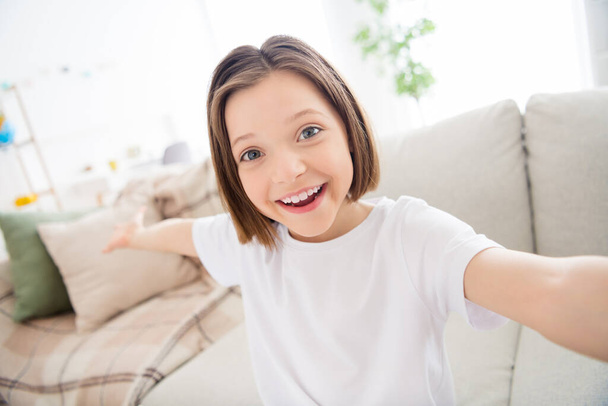 Photo of adorable sweet school girl wear white t-shirt smiling recording video inviting you indoors house home room - Фото, изображение