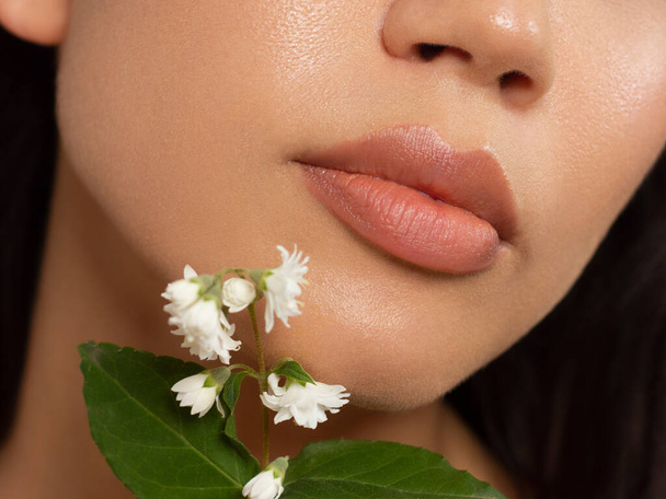 Macro photo of female lips with beautiful skin and nude makeup. light transparent gloss on full lips and white flowers. Fashion photography. Cosmetology and spa. Freshness of spring - Fotografie, Obrázek