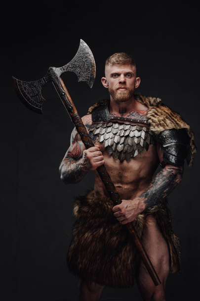 Brutal tattooed warrior wearing light armour and fur holding two-handed axe in dark studio - Photo, Image