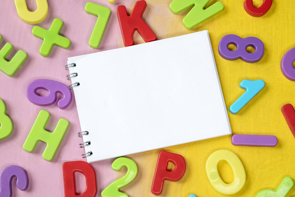A notebook with a white blank sheet on a colorful background among colorful plastic numbers and letters. Educational concept - Photo, Image