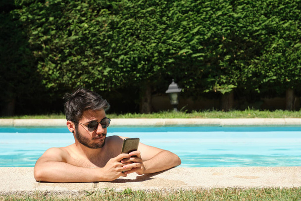 Joven con gafas de sol, usando un smartphone en una piscina. Concepto de verano. - Foto, imagen