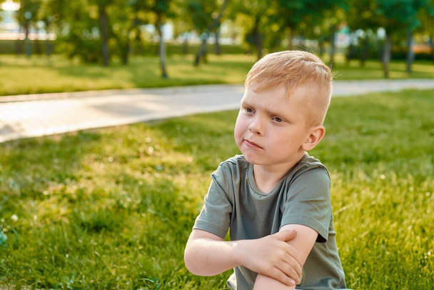 Egy kis ötéves vörös hajú fiú, könyökével a kezében és gondolkodik a parkban, közelről. - Fotó, kép