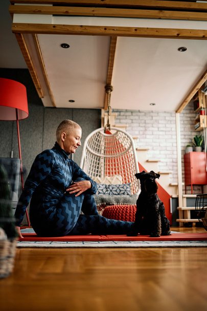 Ελκυστική ηλικιωμένη γυναίκα με κοντά μαλλιά κάνει ασκήσεις γιόγκα στο σπίτι με το σκύλο της. - Φωτογραφία, εικόνα