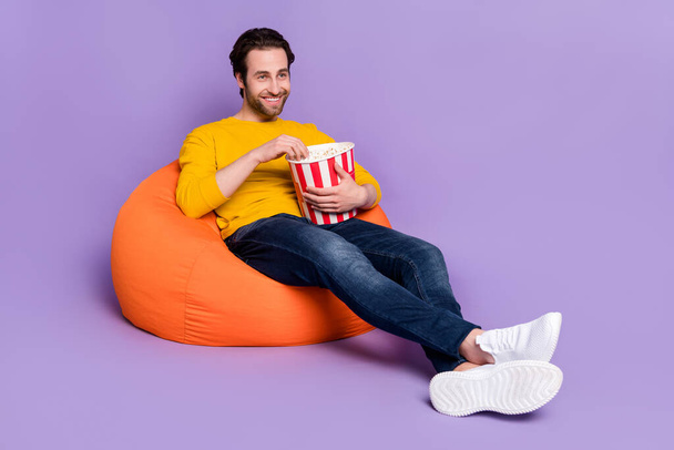 Portrait of attractive cheerful man sitting in bag chair having fun eating corn watching tv isolated over pastel violet purple color background - Fotó, kép