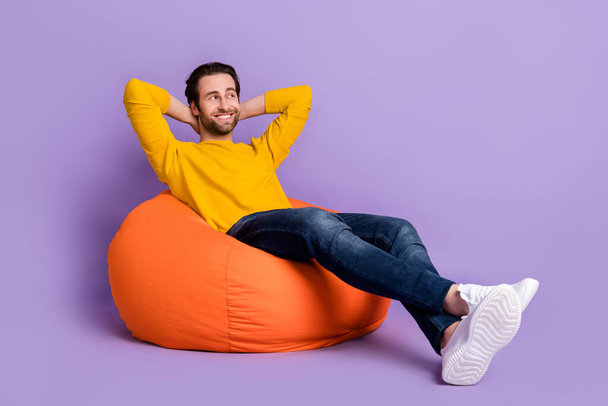 Full length photo of cheerful happy relaxed young man sit bean bag look empty space isolated on purple color background - Фото, зображення