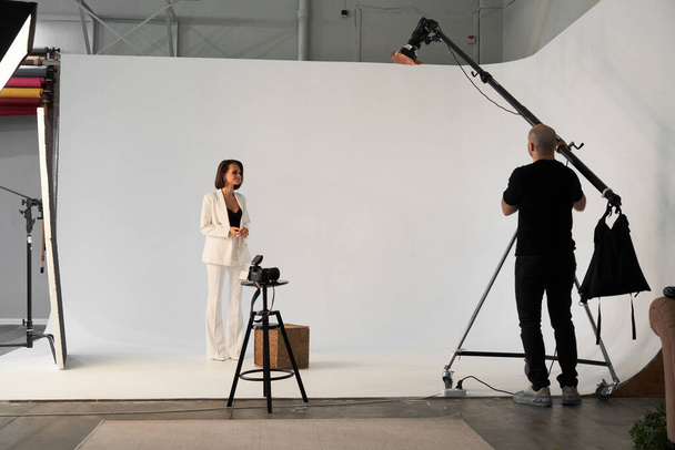 Fashion photography in a photo studio. Professional male photographer taking pictures of beautiful woman model on camera, backstage - Фото, изображение