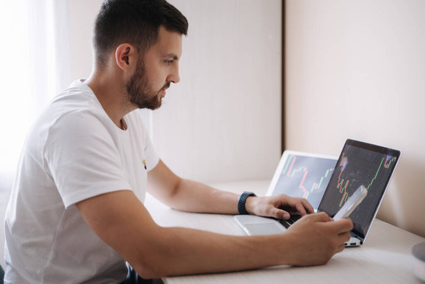 Man looking at finance chart on laptop and tablet, day trader, investing from home. O jovem monitora seus investimentos. Empresário ambicioso - Foto, Imagem