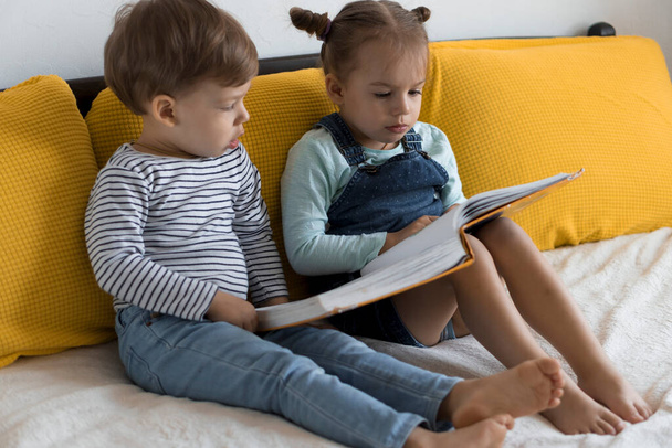 Two preshool, toddler children read large interesting book of fairy tales on yellow bed. Siblings little twins reader have fun, happy kid on quarantine at home. Friendship, family, education concept - Photo, Image