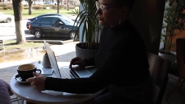 Mladá dívka s brýlemi, krátkými vlasy a piercing v nose sedí v kavárně a pracuje na notebooku. Koncept práce na volné noze a práce na dálku nebo školení. - Záběry, video